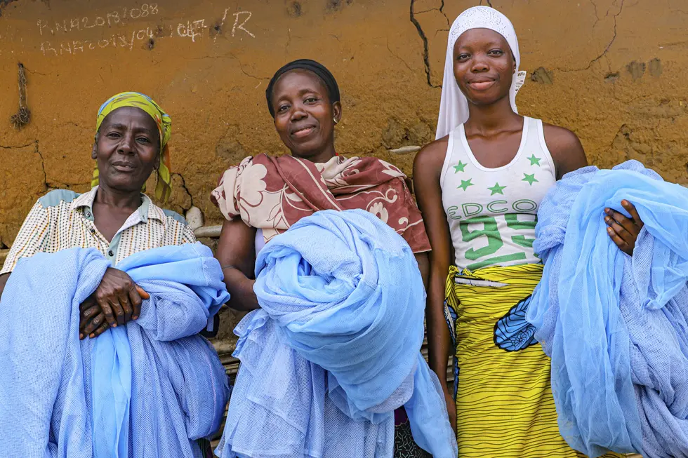 Three women holding bed nets in front of an earthen wall
